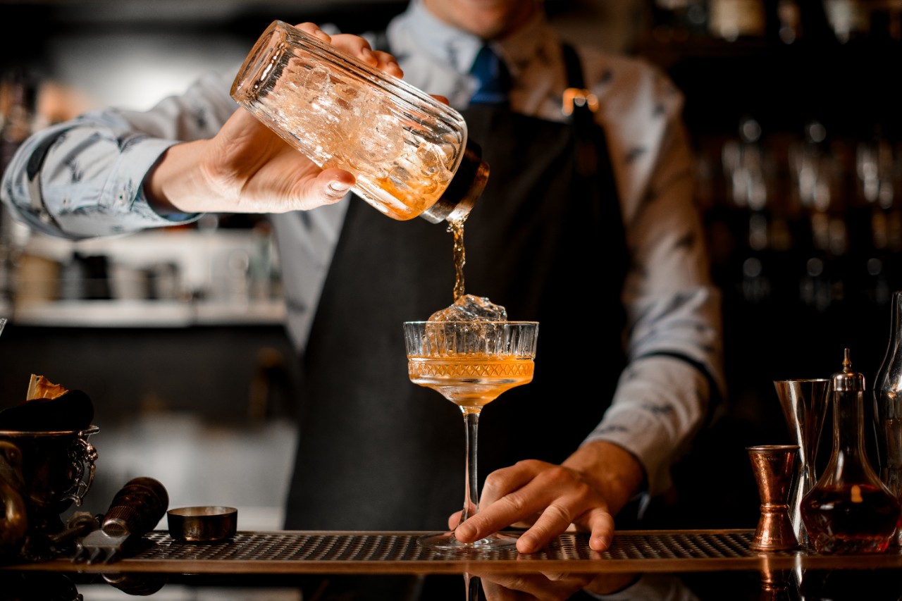 barman gently pours finished cocktail from glass shaker into glass. Body of bartender in black apron on background.
